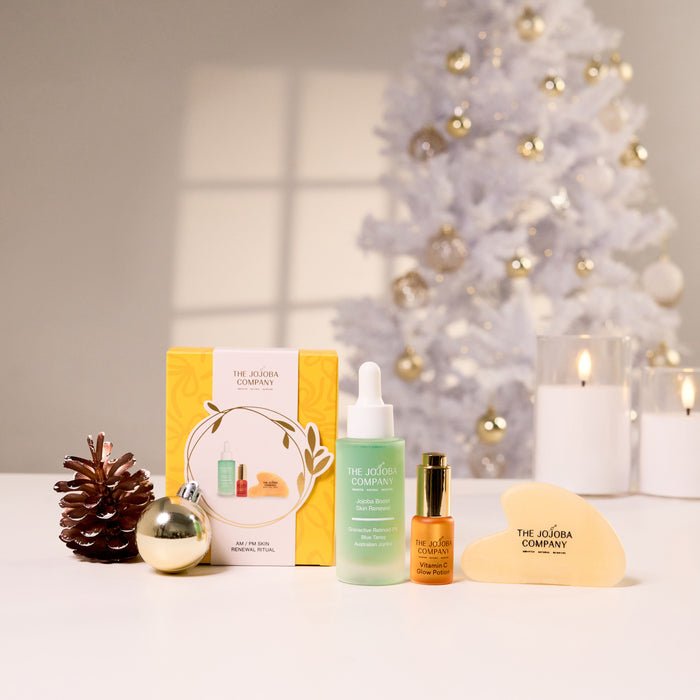 A holiday-themed skincare set from The Jojoba Company arranged on a white surface, featuring a yellow gift box, Jojoba Boost Skin Renewal, Vitamin C Glow Potion, Gua Sha and a gold ornament, with a blurred white Christmas tree and candles in the background.