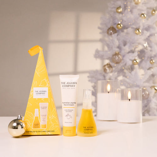 The Jojoba Company Australian Jujube Hand Cream gift set on a white table, featuring a yellow cone-shaped box, a white hand cream tube, a yellow spray bottle, and a gold ornament, styled with white candles and a white Christmas tree in the background.
