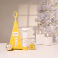 The Jojoba Company Christmas gift set on a white surface, featuring a yellow triangular box, a tube of Australian Jojoba Hand Cream, a tin of Australian Jojoba Lip Balm, and a silver ornament, with a blurred white Christmas tree in the background.