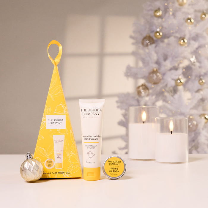 The Jojoba Company Christmas gift set on a white surface, featuring a yellow triangular box, a tube of Australian Jojoba Hand Cream, a tin of Australian Jojoba Lip Balm, and a silver ornament, with a blurred white Christmas tree in the background.