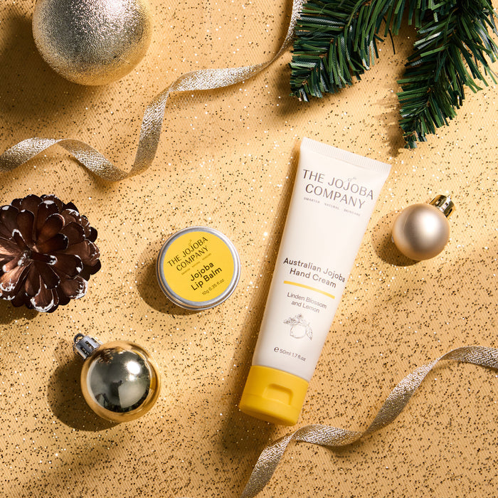 A styled flat-lay of The Jojoba Company products on a glittery gold background with holiday decorations. The products include a tube of Australian Jojoba Hand Cream and a tin of Jojoba Lip Balm.