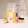 The Jojoba Company holiday gift set arranged on a white surface, featuring a yellow box, a pink-toned bottle with a gold cap, a makeup brush in a gold case, and decorative gold ornaments, with a softly blurred white Christmas tree and candles in the background.
