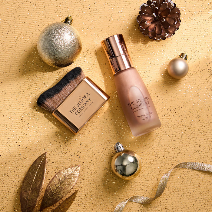 A flat-lay of a beauty product set on a glittery gold background, featuring a bottle of The Jojoba Company Illuminating Jojoba and a matching makeup brush in a gold compact, surrounded by festive gold ornaments and leaves.