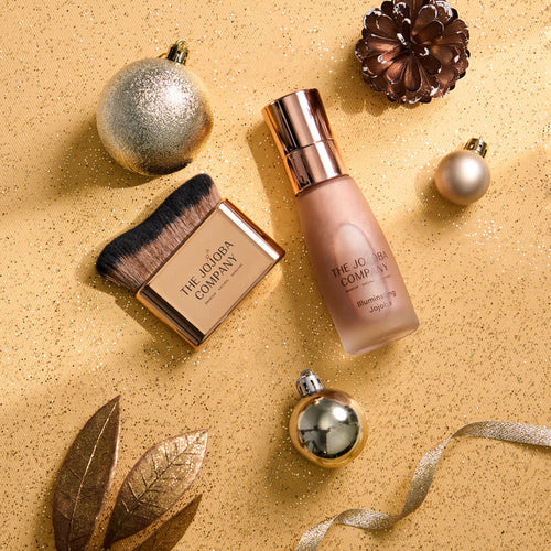 A flat-lay of a beauty product set on a glittery gold background, featuring a bottle of The Jojoba Company Illuminating Jojoba and a matching makeup brush in a gold compact, surrounded by festive gold ornaments and leaves.