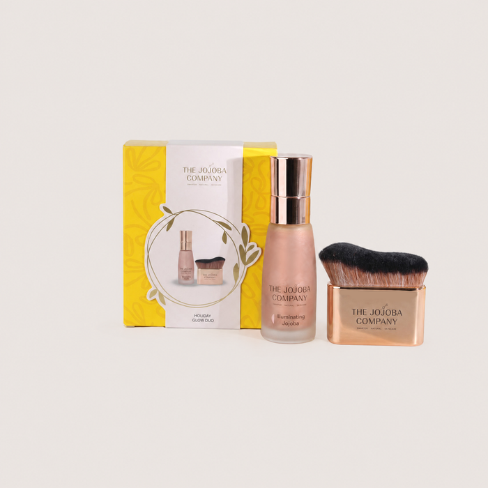 The Jojoba Company Holiday Glow Face set featuring a yellow and white gift box, a frosted bottle of glow serum with a gold cap, and a rose gold makeup brush with black bristles, all arranged on a light beige background.