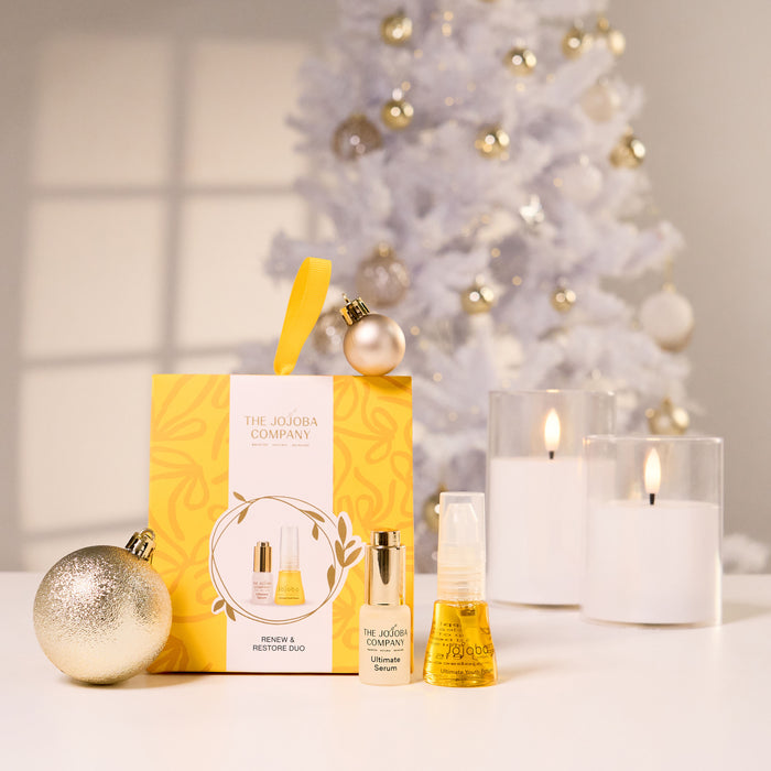 The Jojoba Company Christmas & New Year gift set on a white surface, featuring a yellow gift box with a yellow ribbon, a perfume bottle, a body mist spray, and a scent bag, styled with gold ornaments and white candles in front of a blurred white Christmas tree.