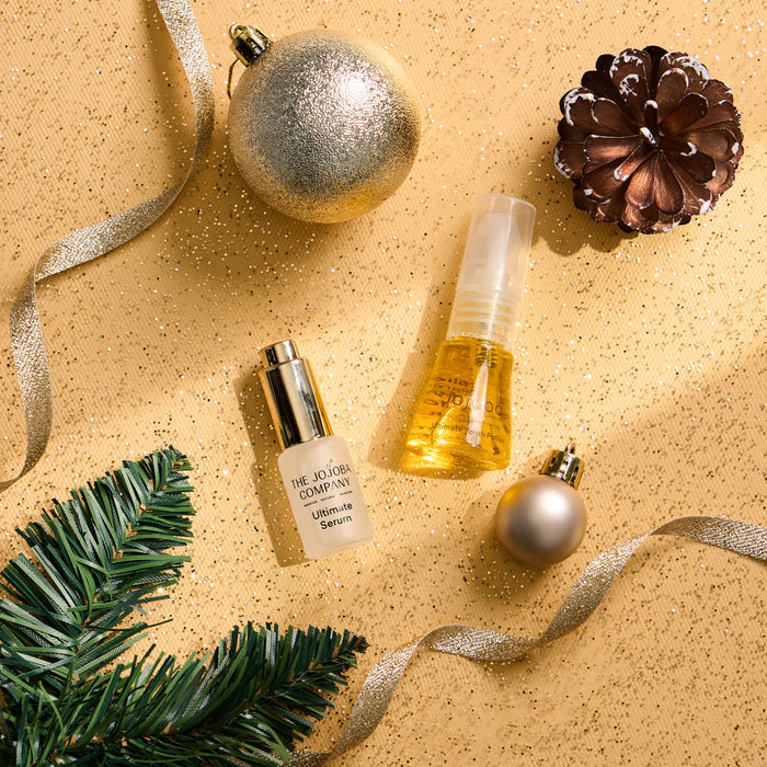 Two cosmetic travel-sized bottles arranged on a glittery gold surface with holiday props, including a silver ornament, pine branch, brown pinecone, and silver ribbon. One bottle is frosted white with a gold cap and reads The Jojoba Company Ultimate Serum