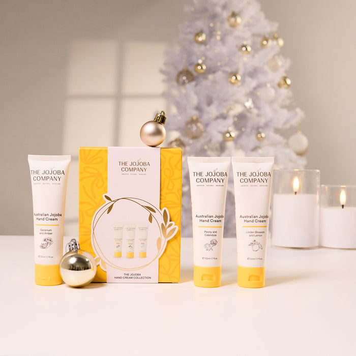 Three tubes of The Jojoba Company Australian Apple Hand Cream arranged in front of a yellow gift box, with decorative gold ornaments and a softly lit white Christmas tree and candles in the background.
