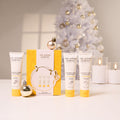 Three tubes of The Jojoba Company Australian Apple Hand Cream arranged in front of a yellow gift box, with decorative gold ornaments and a softly lit white Christmas tree and candles in the background.