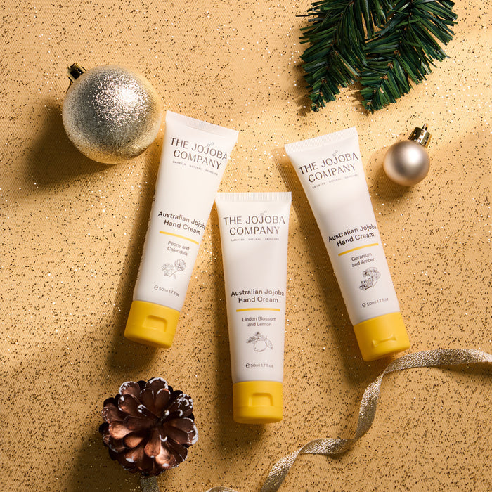 Three tubes of The Jojoba Company Australian Jojoba Hand Cream arranged on a textured gold background with holiday decorations including a silver ornament, pine branch, pinecone, and ribbon.