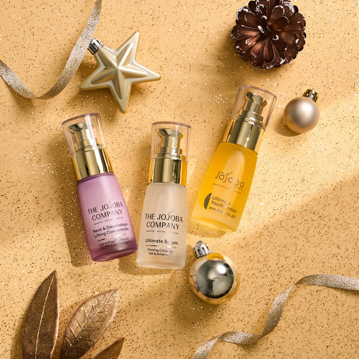Three skincare bottles from The Jojoba Company arranged on a glittery gold surface with holiday decorations. The bottles contain purple, clear, and yellow liquids and have gold pumps.
