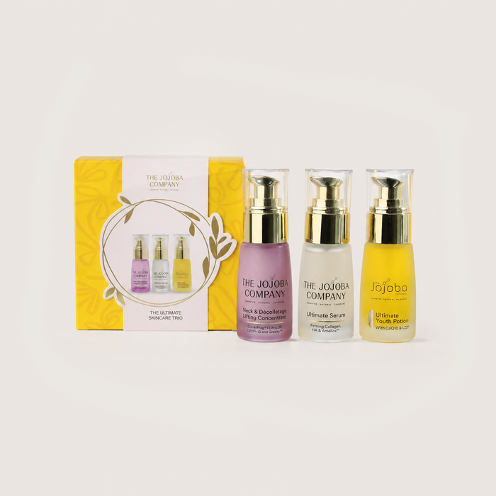 Three skincare bottles from The Jojoba Company with gold pump caps are displayed in front of their yellow and white packaging box on a light beige background.