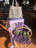 canvas tote bags with handprinted  the Lavender By The Bay logo hanged on a rack behined a blue chair with lavender flowers in a wooden floor store