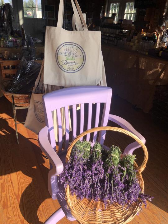 canvas tote bags with handprinted  the Lavender By The Bay logo hanged on a rack behined a blue chair with lavender flowers in a wooden floor store