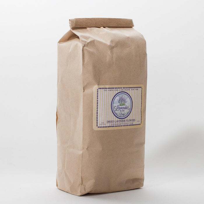 A closed brown paper bag filled with dried French loose lavender buds. The bag has a label that says "Lavender by the Bay" and "Dried Lavender Flowers. shot taken  at an angle