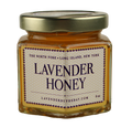 A hexagonal glass jar filled with golden raw honey, with a gold lid. The jar has a label reading "Lavender Honey" and the location "The North Fork, Long Island, New York