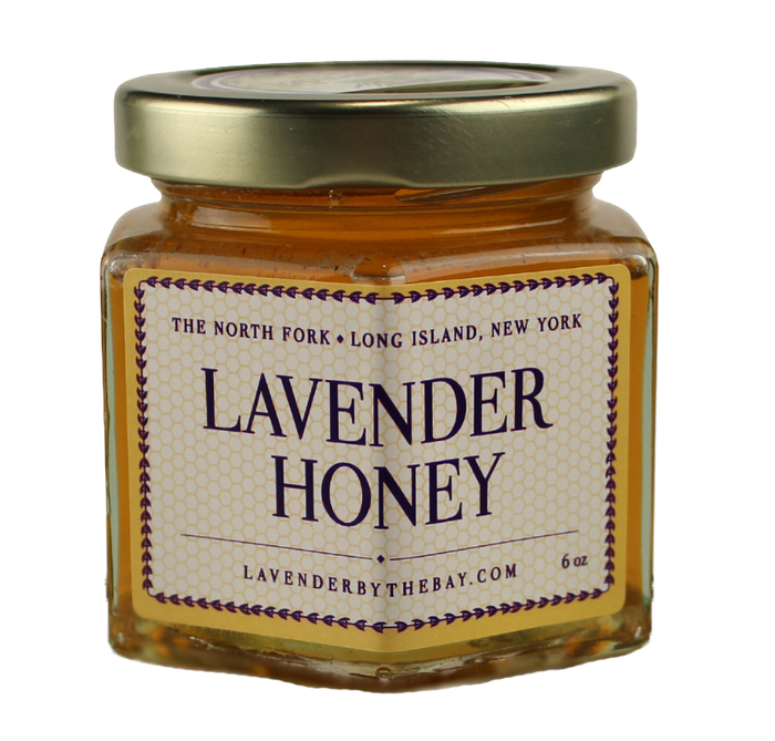 A hexagonal glass jar filled with golden raw honey, with a gold lid. The jar has a label reading "Lavender Honey" and the location "The North Fork, Long Island, New York