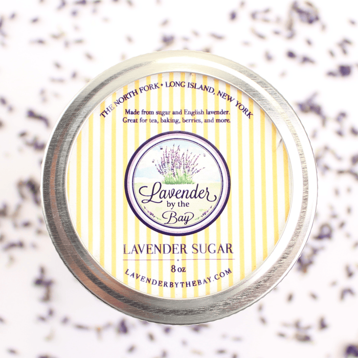 A round metal jar filled with lavender sugar, with a colorful label that includes the name "Lavender by the Bay" and information "made from sugar and english lavender great for tea, baking, barries and more"