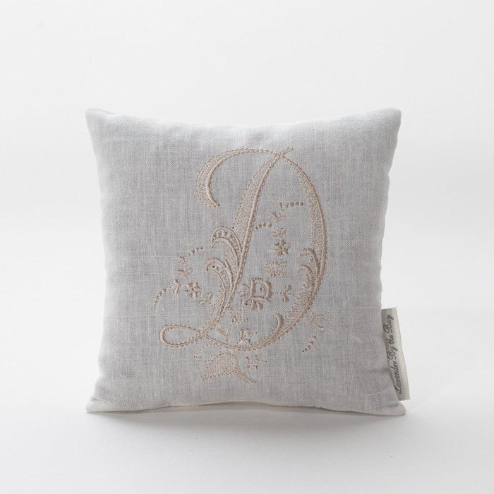 The image depicts a beautifully square, off-white embroidered monogram pillow with the letter "D," intricately stitched with delicate floral patterns and swirls. The pillow appears soft and luxurious.