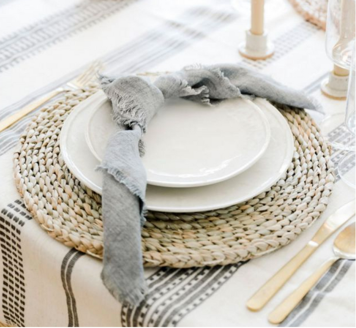 Dining table setting with white plates, gray Ethiopian cotton napkin, and a woven plate charger on a hand-woven tablecloth.
